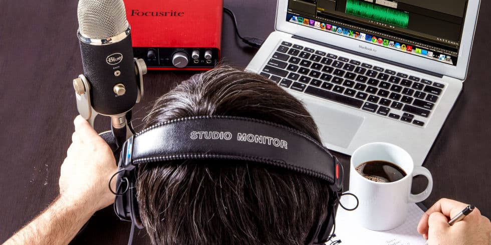 Image shows an photo taken from above a man, who is sat at a home studio. The man is wearing headphones, and on the desk is a micrphone, an audio interface, a laptop and a cup of coffee.