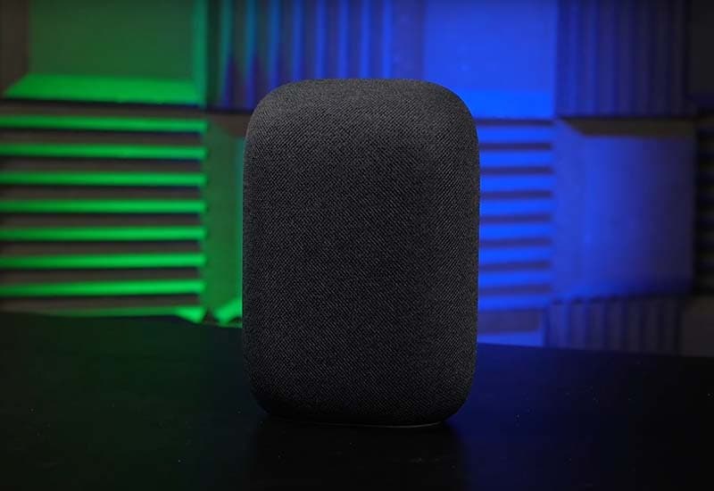 Image shows a grey Google Nest smart speaker, placed upright on a table with green and blue lights highlighting the wall behind it.