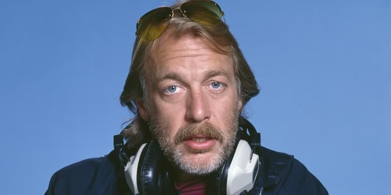 Image shows a still from the TV show, WKRP in Cincinnati. Image shows a white, middle-aged man from the shoulders up, Johnny Fever, looking directly at the camera against a blue background..