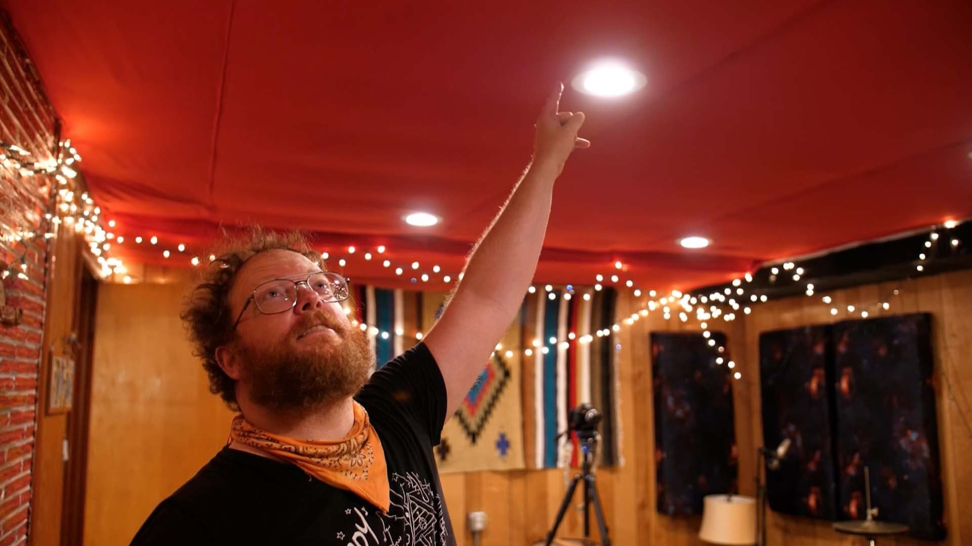 A white man in the Shady Pines radio booth, points and looks upwards to the ceiling that has pillows for acoustic treatment.