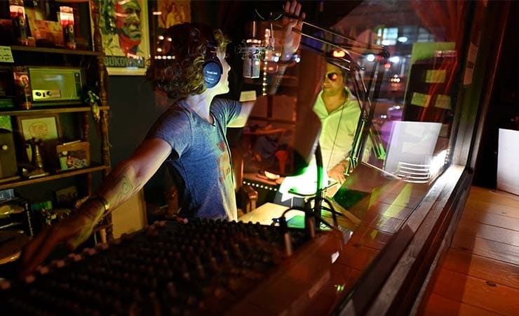 A woman presenting a radio show and talking to a guest, behind a glass screen.