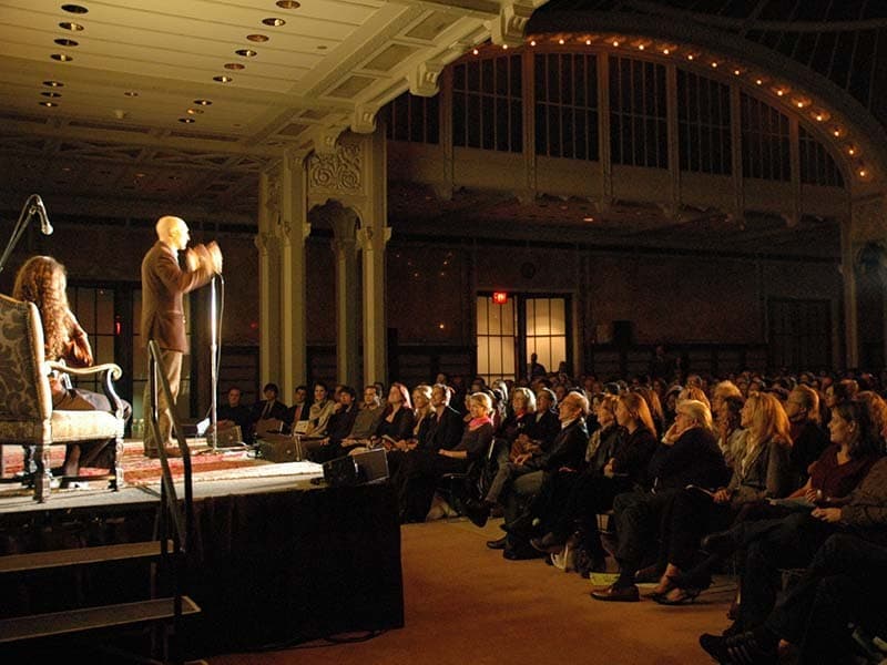 Image shows a man standing on stage, speaking in to a microphone and to a large audience.