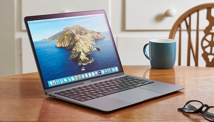 A macbook on a kitchen table, with a pair of glasses and coffee cup next to it.